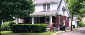 Exterior of two story house with large covered front porch, red brick, and white cedar shake shingles