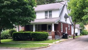 Exterior of two story house with large covered front porch, red brick, and white cedar shake shingles