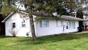 Exterior of ranch house with small lawn, light vertical siding, and pale blue shutters.