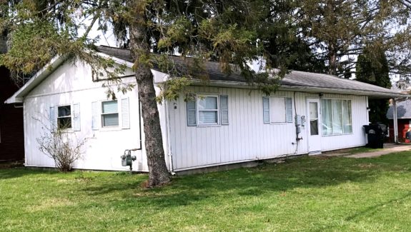 Exterior of ranch house with small lawn, light vertical siding, and pale blue shutters.