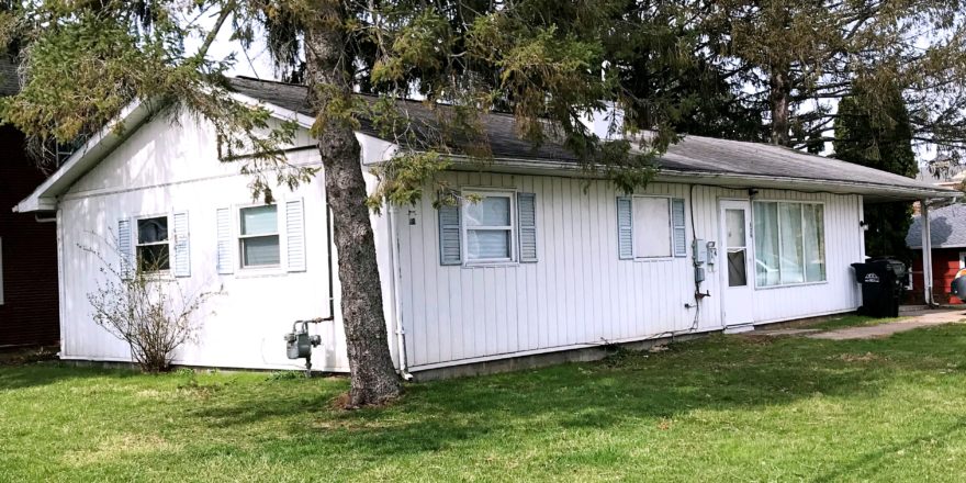 Exterior of ranch house with small lawn, light vertical siding, and pale blue shutters.