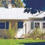 Exterior of small, one-story home with light siding and green shutters