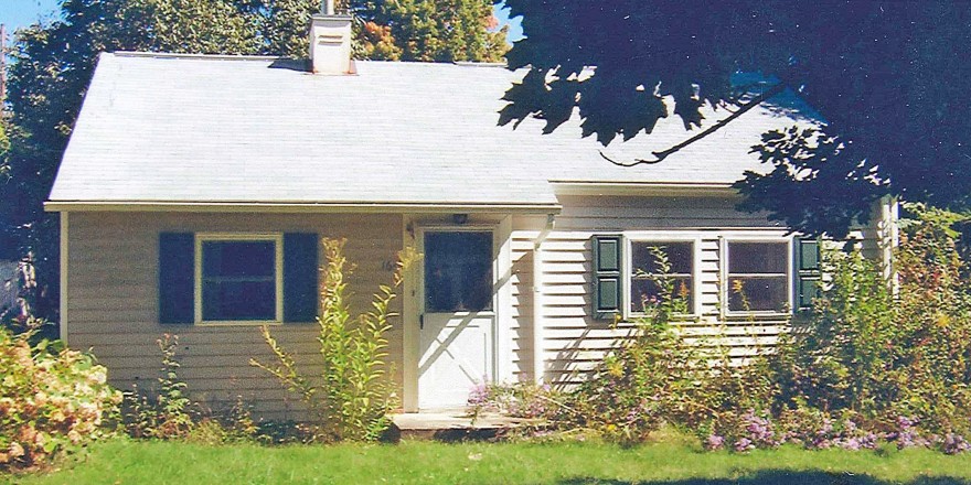 Exterior of small, one-story home with light siding and green shutters