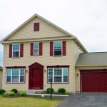 Exterior of two story house with large garage, tan siding and maroon front door, shutters, and garage door