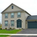 Exterior of house with tan siding, green front door, garage door, and trim, driveway, and lawn