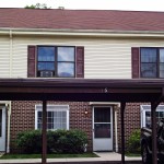Exterior of townhouse with brick first floor and yellow siding with maroon shutters on the second floor with carport out front