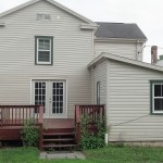 Back exterior of house with tan siding and green trim. Red wood deck attached to back french doors