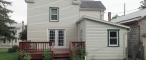 Back exterior of house with tan siding and green trim. Red wood deck attached to back french doors