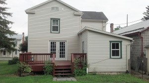Back exterior of house with tan siding and green trim. Red wood deck attached to back french doors