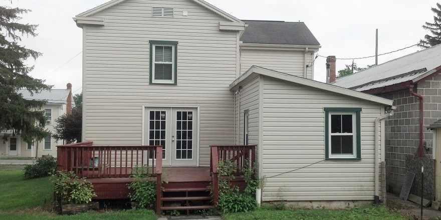 Back exterior of house with tan siding and green trim. Red wood deck attached to back french doors