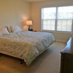 Carpeted bedroom with queen bed, dresser, chair, nightstand with lamp, and large window
