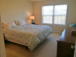Carpeted bedroom with queen bed, dresser, chair, nightstand with lamp, and large window