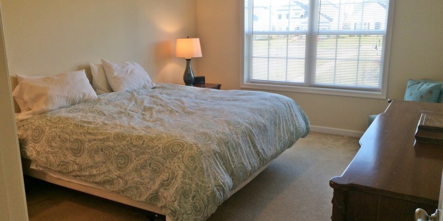 Carpeted bedroom with queen bed, dresser, chair, nightstand with lamp, and large window