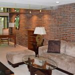 Living room with exposed brick wall; brown, suede couch, glass accent tables, and wooden desk and bookshelf