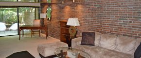 Living room with exposed brick wall; brown, suede couch, glass accent tables, and wooden desk and bookshelf