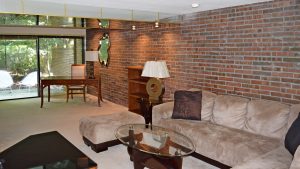 Living room with exposed brick wall; brown, suede couch, glass accent tables, and wooden desk and bookshelf