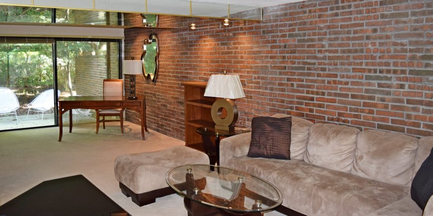 Living room with exposed brick wall; brown, suede couch, glass accent tables, and wooden desk and bookshelf