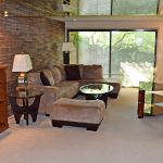 Living room with exposed brick wall; brown, suede couch, glass accent tables, and wooden desk and bookshelf