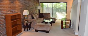 Living room with exposed brick wall; brown, suede couch, glass accent tables, and wooden desk and bookshelf