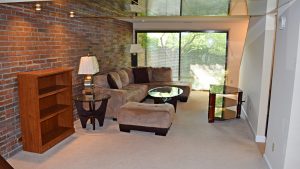 Living room with exposed brick wall; brown, suede couch, glass accent tables, and wooden desk and bookshelf