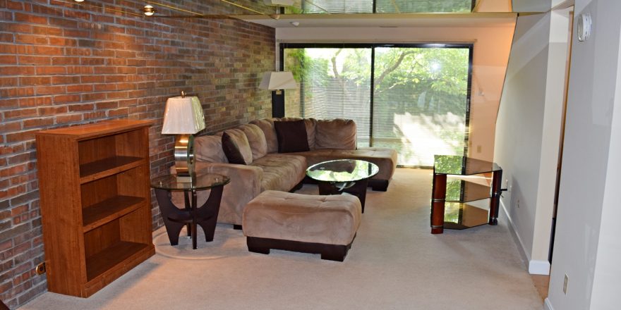 Living room with exposed brick wall; brown, suede couch, glass accent tables, and wooden desk and bookshelf