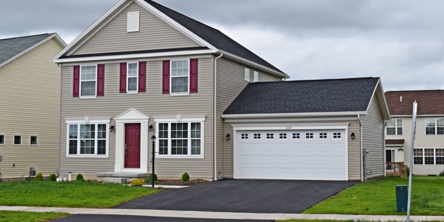Exterior of house with tan siding, red front door, white garage door and trim, driveway, and lawn