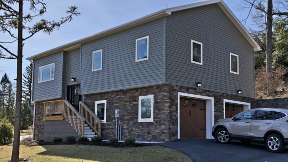 House with gray siding and stone siding. Stairs lead up to the front door and there are two garage doors on the side.