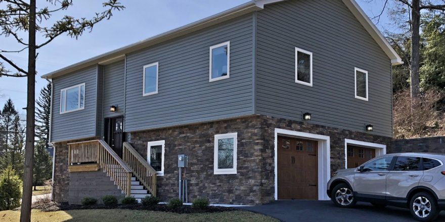 House with gray siding and stone siding. Stairs lead up to the front door and there are two garage doors on the side.