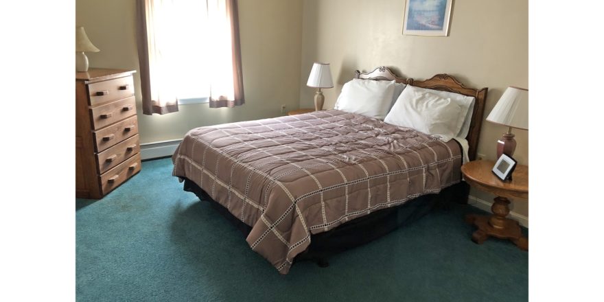 Carpeted bedroom with queen bed, two end tables with lamps, picture, window, and dresser with five drawers and a lamp on top