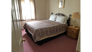 Carpeted bedroom with queen bed, window, picture, and wicker nightstand with lampe