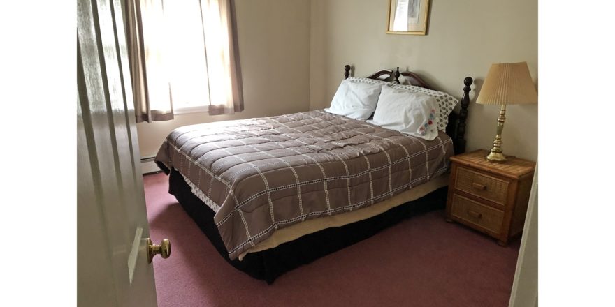 Carpeted bedroom with queen bed, window, picture, and wicker nightstand with lampe