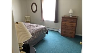 Carpeted bedroom with queen bed, window, mirror, chair, nightstand with lamp, and wooden dresser with five drawers and lamp