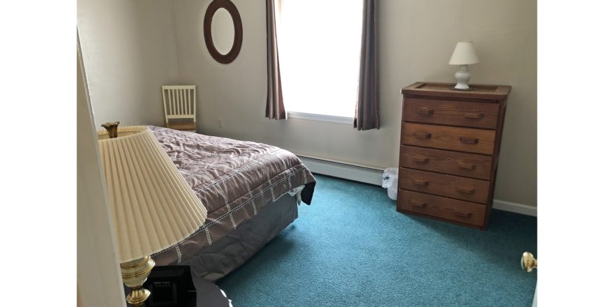 Carpeted bedroom with queen bed, window, mirror, chair, nightstand with lamp, and wooden dresser with five drawers and lamp