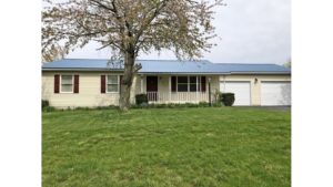 Exterior of ranch house with yellow siding and maroon shutters, covered front porch, two car garage, and tree