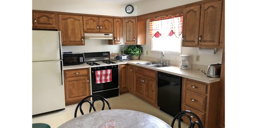 Kitchen with white and black appliances, wood cabinets, table with chairs and stool, and small window above the sink