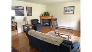 Living room with corner fireplace, pictures, couch and armchair, coffee table, end table, rocking chair, and bookcase