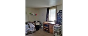 Carpeted bedroom with twin bed, lamp, window, desk and shelving