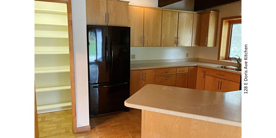 Kitchen with large pantry, black fridge, light wood cabinets, parkay flooring, and stone countertops.