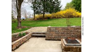 Sunken patio with fire pit. Lawn with trees and flower bushes.