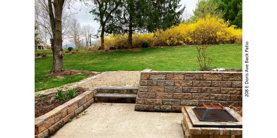 Sunken patio with fire pit. Lawn with trees and flower bushes.