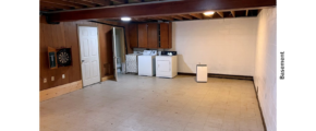 a large basement with white walls, white tile, wood beams on the ceiling, and a washer and dryer unit