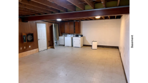 a large basement with white walls, white tile, wood beams on the ceiling, and a washer and dryer unit