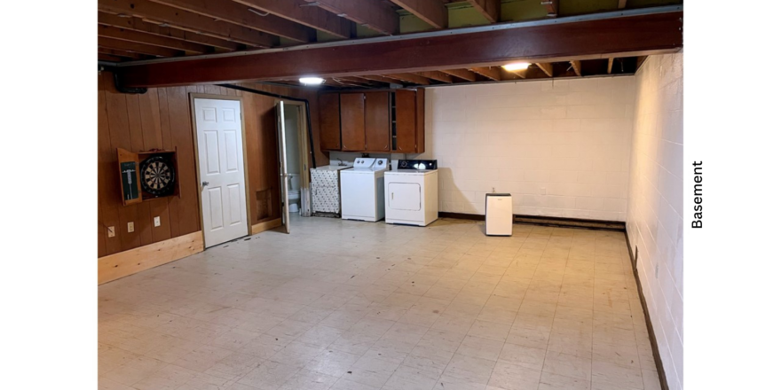 a large basement with white walls, white tile, wood beams on the ceiling, and a washer and dryer unit