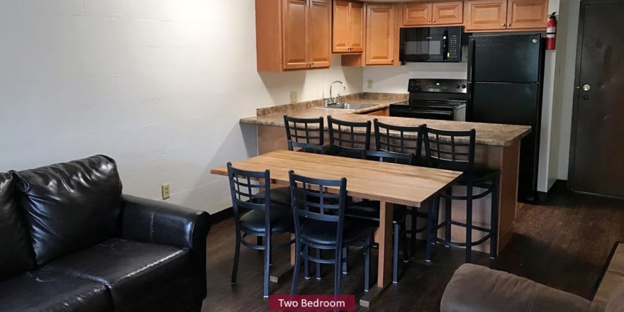 Kitchen with black appliances, wood-tone cabinets and bar seating. Living Room with couch and dining table.