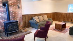Carpeted basement den with wood wainscot, brick hearth, wood burning fireplace, and furniture