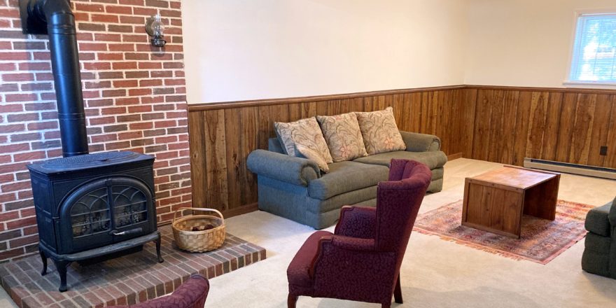 Carpeted basement den with wood wainscot, brick hearth, wood burning fireplace, and furniture