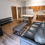 Kitchen with wood-tone cabinets and white appliances, island, and living room with sofa, loveseat, coffee table and TV stand
