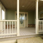 Main entry covered front porch with cream colored railings and hardwood decking. Large ornate front door with a storm door and windows flanking each side