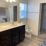 a bathroom with a white toilet, double sink with dark brown cabinets and drawers, a large mirror, and a closet