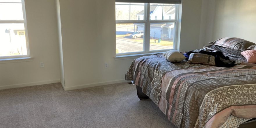 a bedroom with beige carpet, a bed, and two windows